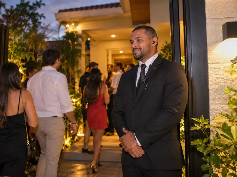 Security guard in a black suit and earpiece welcoming guests at the lit entrance of a party