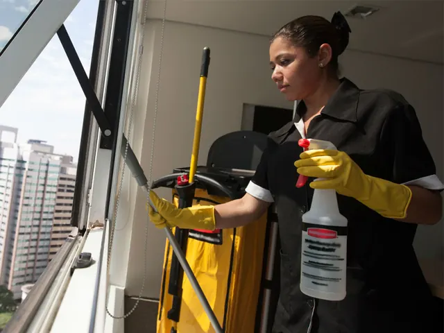 Cleaner in uniform and yellow gloves cleaning an apartment window next to a cleaning cart