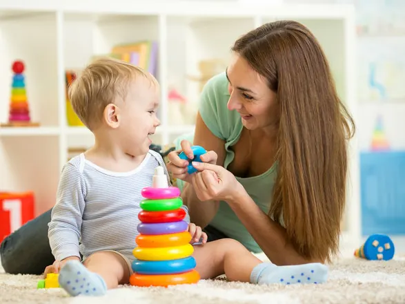 Infant nanny smiling at a baby playing with colorful stacking rings on the bedroom rug